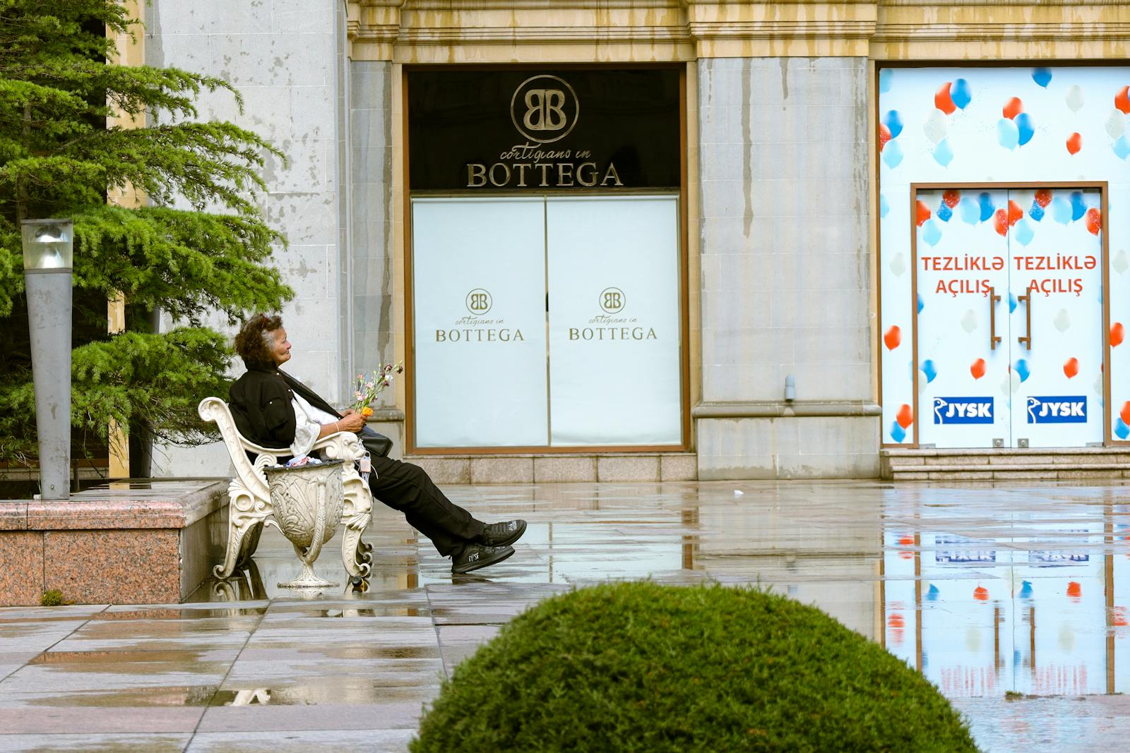 A man holding a bouquet sits on a bench outside shops in Baku, Azerbaijan.